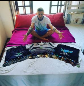 Foto do Márcio sentando na cama com dezenas de medalhas, camisetas dos seus apoiadores(X One Academia, Levi Personal Trainer e Aflodef)