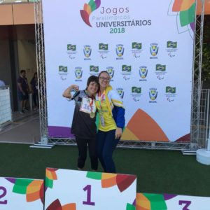 Foto da Kamila e da professora Bruna nas Paralímpiadas Universitárias, com sua medalha no pódio. 