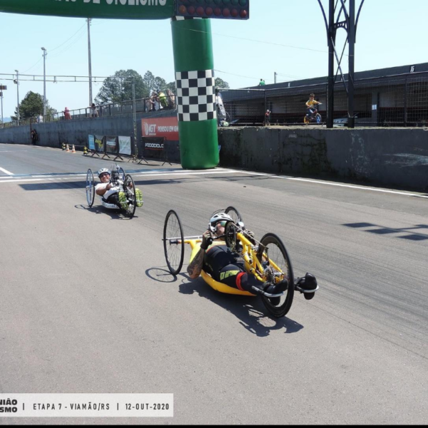 Foto do Rodrigo Schu na chegada da ultima competição que ele participou(16º Copa União de Ciclismo - Etapa 7 - Viamão-RS). Ele está com sua handbike amarela e ao fundo esta mais um atleta chegando e o portico de chegada inflave na cor verde.