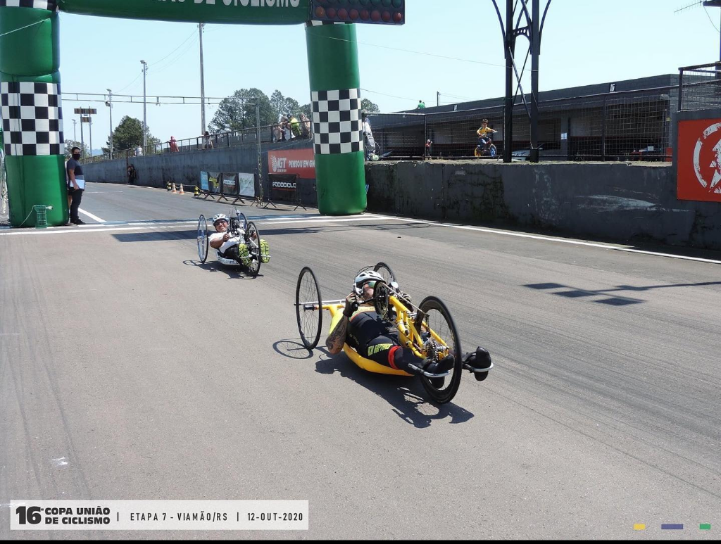 Foto do Rodrigo Schu na chegada da ultima competição que ele participou(16º Copa União de Ciclismo - Etapa 7 - Viamão-RS). Ele está com sua handbike amarela e ao fundo esta mais um atleta chegando e o portico de chegada inflave na cor verde.